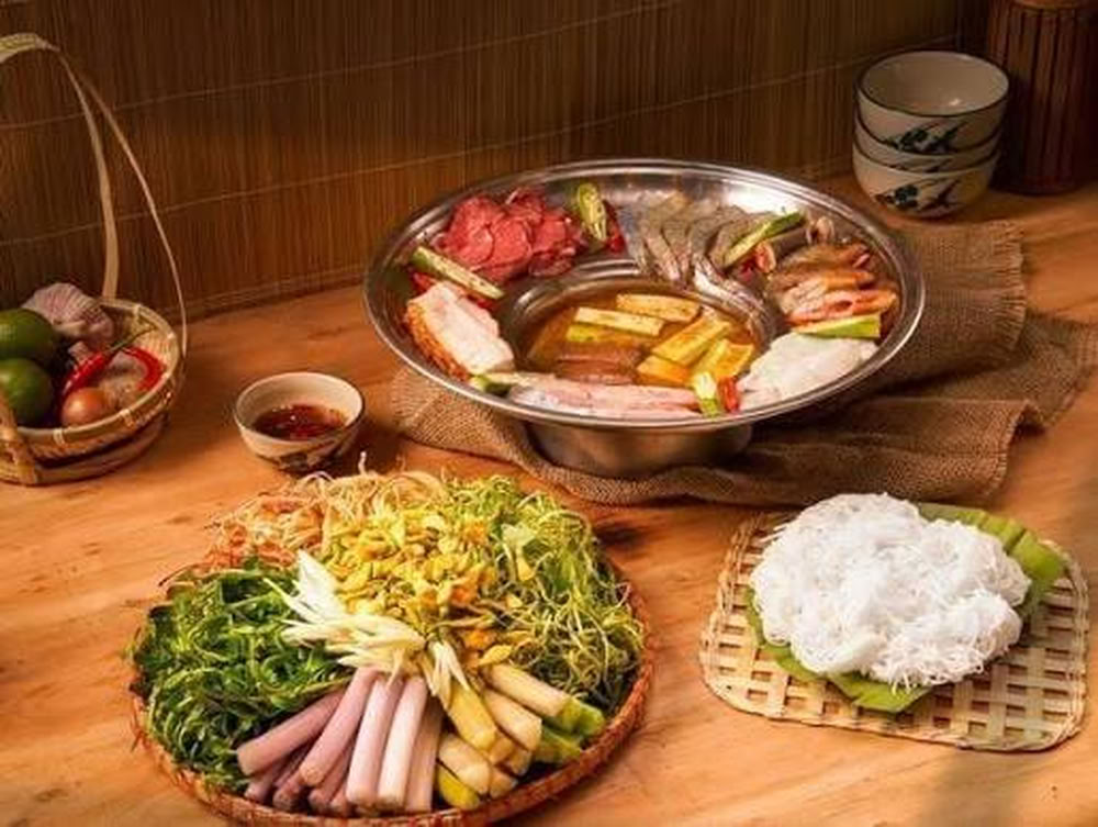 Une table dressée avec une fondue de poisson linh, accompagnée de fleurs de sesbanie jaune, de légumes du Mékong et de vermicelles de riz.